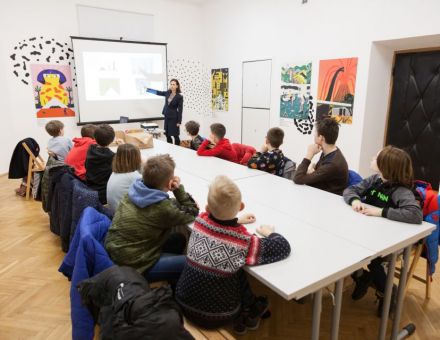 Warsztat architektoniczny [warsztat dla dzieci w ramach Zimowej Majsterni. Na zdjęciu widzimy dzieci siedzące przy stole, słuchające Pani prowadzącej. Tłumaczy ona zagadnienia związane z tematem architektury, pokazując na obraz wyświetlony przez projektor]