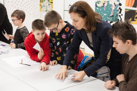 Warsztat architektoniczny  [warsztat dla dzieci w ramach Zimowej Majsterni. Zbliżenie na dzieci i prowadzącą, którzy znajdują się przy stole. Prowadząca przykłada wycięte szkice budynków do stołu, a dzieci przyglądają się ze skupieniem]