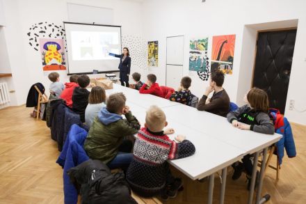 Warsztat architektoniczny [warsztat dla dzieci w ramach Zimowej Majsterni. Na zdjęciu widzimy dzieci siedzące przy stole, słuchające Pani prowadzącej. Tłumaczy ona zagadnienia związane z tematem architektury, pokazując na obraz wyświetlony przez projektor]