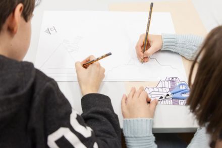 Warsztat architektoniczny [warsztat dla dzieci w ramach Zimowej Majsterni. Na zdjęciu chłopiec i dziewczynka odrysowują przykładowe szkice budynków, które leżą obok kartki]