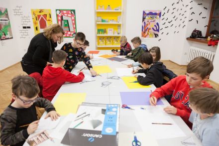 Warsztat architektoniczny n [warsztat dla dzieci w ramach Zimowej Majsterni. Na zdjęciu widzimy dzieci siedzące przy stole, wykonujące prace z zakresu architektury. Konsultują się z prowadzącą. Odrysowują budynki, pracują w parach]