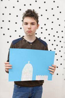 Warsztat architektoniczny warsztat dla dzieci w ramach Zimowej Majsterni. Zdjęcie przedstawia chłopca trzymającego makietę miasta. Praca w kolorach niebiesko-białym. Znajduje się na niej  kaplica oraz szereg niewielkich budynków]