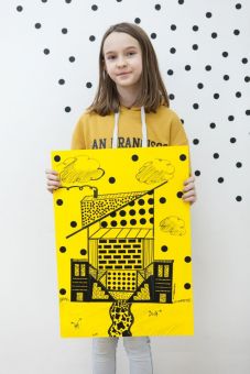 Na zdjęciu stoi dziewczynka, trzymająca swój plakat utrzymany w kolorystyce żółto-czarnej. Przedstawia budynek pośród natury.