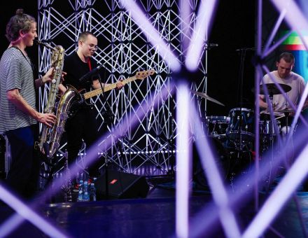 Trzech muzyków: saksofonista, gitarzysta i perkusista. Zdjęcie zrobione spomiędzy aluminiowych kratownic, które otaczają grupę.