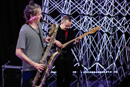 Dwóch muzyków, jeden z saksofonem, drugi z gitarą elektryczną. Obaj grają na instrumentach, są na nich skupieni. W tle aluminiowe kratownice.