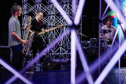 Trzech muzyków: saksofonista, gitarzysta i perkusista. Zdjęcie zrobione spomiędzy aluminiowych kratownic, które otaczają grupę.