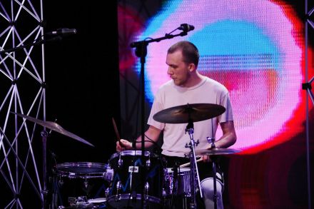 Perkusista gra na swoim instrumencie. W tle na ekranie led za jego  plecami widać animację z różowym kołem.