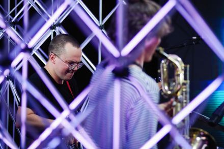 Zdjęcie zrobione spomiędzy aluminiowych kratownic, które otaczają grupę. Zbliżenie na twarz gitarzysty, ma zaciśnięte usta, daje z siebie wszystko. Obok odwrócony plecami do aparatu saksofonista.