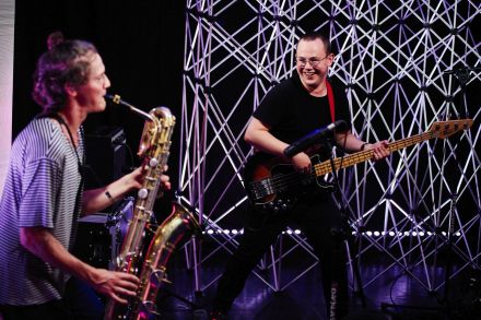 Saksofonista i gitarzysta grają na swoich instrumentach, gitarzysta śmieje się i patrzy na saksofonistę. W tle aluminiowe kratownice/