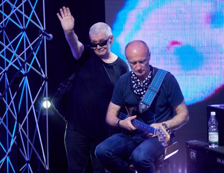 Wokalista stoi i ma podniesioną do góry rękę, macha – pozdrawia. Obok na stołku siedzi gitarzysta, jest skupiony na instrumencie. Za plecami obu mężczyzn ekran led z abstrakcyjną animacją w kolorze błękitnym.