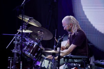 Perkusista gra na swoim instrumencie. W tle na ekranie led za jego plecami widać animację z szarym kołem. Perkusista ma blond dredy.