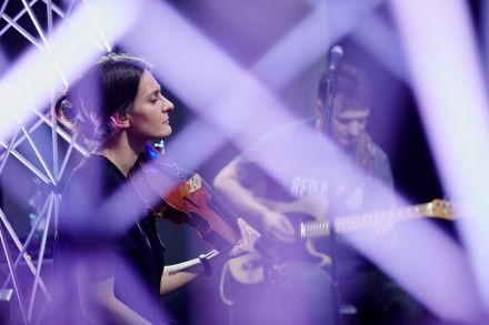 Gitarzysta i skrzypaczka. Kobieta ma zamknięte oczy, wsłuchuje się w muzykę graną przez jej towarzyszy. Skrzypce trzyma na ramieniu w gotowości, ale dłoń ze smyczkiem ma opuszczoną. Zdjęcie zrobione spomiędzy aluminiowych kratownic, które otaczają grupę.