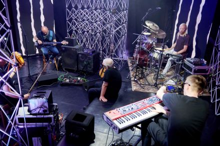 Zdjęcie z góry i z szerszej perspektywy. Na zdjęciu czterech muzyków: gitarzysta, perkusista, klawiszowiec oraz wokalista. Wokół nich ekrany led z szarymi abstrakcyjnymi animacjami oraz aluminiowe kratownice. Na podłodze dużo elektronicznego sprzętu: głośniki, kable, wzmacniacze. Z grupy wyróżnia się wokalista, który siedzi zrelaksowany podłodze i przygląda się grającym muzykom.