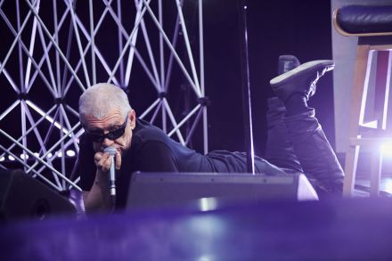 Zbliżenie na wokalistę. Na oczach ma czarne okulary, w ręku trzyma mikrofon. Być może właśnie śpiewa lub mówi, ale nie widać tego, bo mikrofon zasłania usta. Wokalista leży na brzuchu, ma podniesione do góry nogi i macha nimi, jak gdyby leżał na łące pełnej kwiatków.  W tle jednak nadal są aluminiowe kratownice.