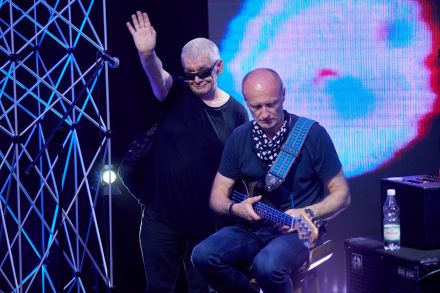 Wokalista stoi i ma podniesioną do góry rękę, macha – pozdrawia. Obok na stołku siedzi gitarzysta, jest skupiony na instrumencie. Za plecami obu mężczyzn ekran led z abstrakcyjną animacją w kolorze błękitnym.