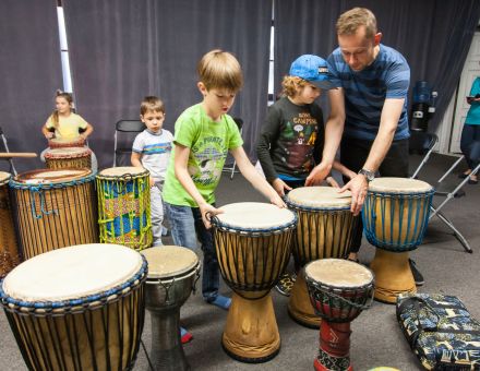 Dwóch chłopców z tatą wybierają sobie bębny. Przed nimi stoi aż sześć do wyboru. Za nimi także kilka. W tle inne dzieci.