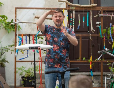 Prowadzący warsztaty pokazuje coś dłońmi. Obok niego stoi elektroniczny instrument muzyczny. W tle kolorowe wietrzne dzwonki zawieszone na drewnianym stelażu.