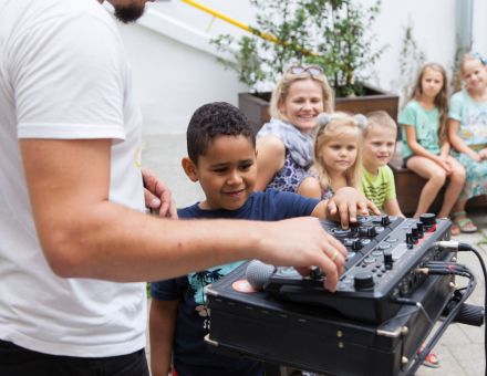Chłopiec sprawdza jak działają suwaki i pokrętła konsolety, wszystko pod czujnym okiem prowadzącego. W tle kolejka dzieci, które także chcą spróbować.