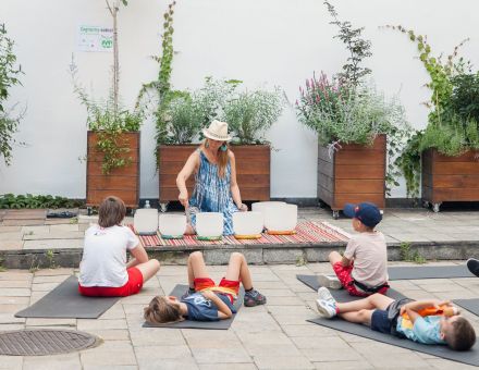 Dzieci leżą na matach na podwórku staromiejskiej kamienicy, przed nimi kobieta grająca na misach.