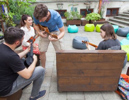 Patio Warsztatów Kultury. Na zdjęciu trzech uczestników w tym mężczyzna, kobieta i nastolatka. Wszyscy siedzą na drewnianych ławkach i trzymają w rękach ukulele. Do mężczyzny podchodzi prowadzący warsztaty, pochyla się nad nim i pokazuje mu jak trzymać chwyt na ukulele.