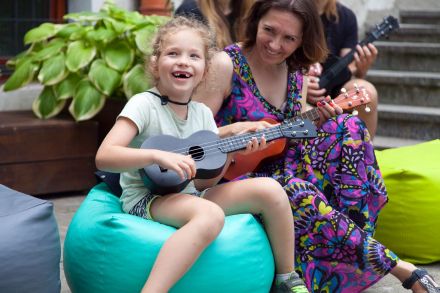 Matka z córką, uwagę przyciąga uśmiech dziewczynki, a szczególnie to, że nie ma dwóch mlecznych jedynek. Jest ubrana na zielono i gra na czarnym ukulele. Obok niech uśmiechnięta matka w fioletowej kwiecistej sukni.