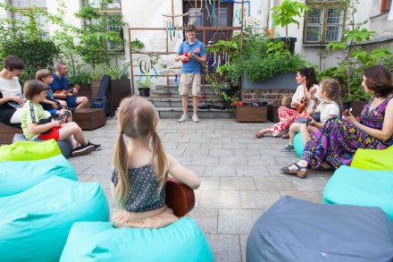 Warsztaty gry na ukulele odbywają się na patio staromiejskiej kamienicy, prowadzący stoi pośrodku, wokół uczestnicy siedzą w kole na workowatych kolorowych pufach, w rękach trzymają instrumenty.
