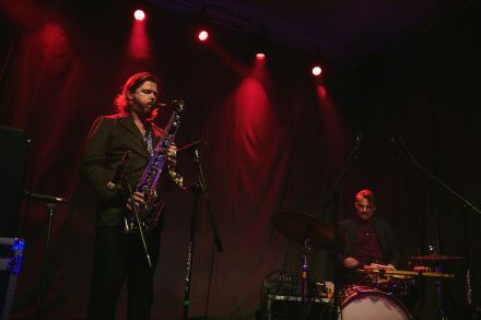 Saksofonista i perkusista, skupieni. Saksofonista ma zamknięte oczy. W tle ciemne kotary oświetlone czerwonym światłem.