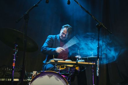 Zbliżenie na perkusistę, ma zaciśnięte oczy i z całej siły uderza pałeczkami w bębny. Wokół unosi się niebieski dym.