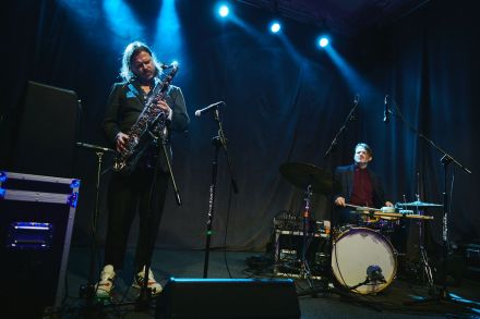 Saksofonista i perkusista, obaj muzycy grają. W tle ciemne kotary oświetlone czerwonym światłem.