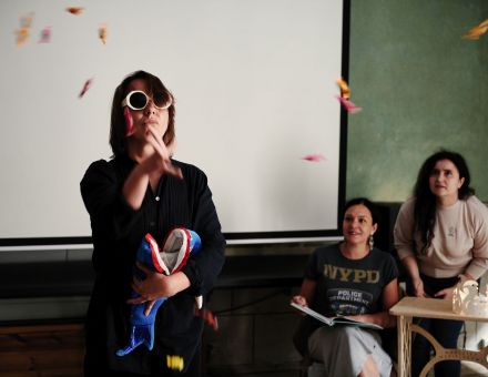 Trzy kobiety znajdują się w pokoju staromiejskiej kamienicy. Na zielonej ścianie wisi ekran do rzutnika. Jedna z kobiet w pierwszym planie. Ma białe przeciwsłoneczne okulary, w rękach trzyma otwarty plecak-wieloryb. Kobieta podrzuciła konfetti, które wokół niej powoli opada na podłogę. Na drugim planie dwie kobiety przyglądają się tej pierwszej, uśmiechają się.