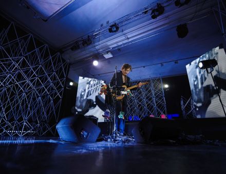 Szeroki plan, sala koncertowa, na suficie szyny z reflektorami, w tle aluminiowe kratownice, ekrany led z abstrakcyjnymi animacjami. Oświetlenie niebieskie. Na środku stoi muzyk, gra na gitarze do której na sznurkach przyczepione są pluszowe maskotki.