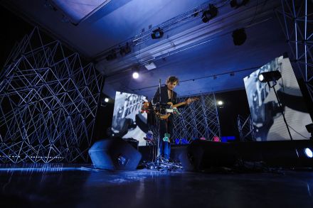 Szeroki plan, sala koncertowa, na suficie szyny z reflektorami, w tle aluminiowe kratownice, ekrany led z abstrakcyjnymi animacjami. Oświetlenie niebieskie. Na środku stoi muzyk, gra na gitarze do której na sznurkach przyczepione są pluszowe maskotki.