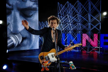 Wokalista w centrum zdjęcia. Ma zamknięte oczy, podniesioną  lewą rękę, drugą ręką trzyma gitarę, szeroko otwarte usta. Śpiewa do mikrofonu. na gitarze na sznurkach przyczepione są pluszowe maskotki. W tle ekran led z jego twarzą oraz aluminiowe kratownice i niebiesko-różowy ledon w kształcie liter INNE.