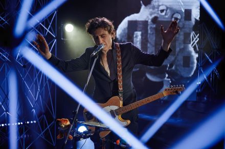 Zdjęcie zrobione zza aluminiowych kratownic. Wokalista na środku. Obie ręce podniesione do góry, gitara trzyma się tylko na pasku. Mężczyzna ma zamknięte oczy i śpiewa do mikrofonu.