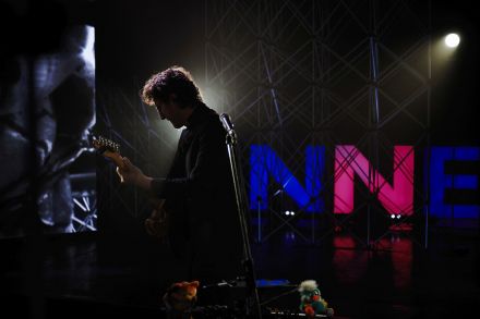 Muzyk stoi bokiem do obiektywu aparatu, gra na gitarze, w tle aluminiowe krafownice i niebiesko-różowy leon w kształcie liter INNE. Zdjęcie jest dość mroczne, mało na nim światła.