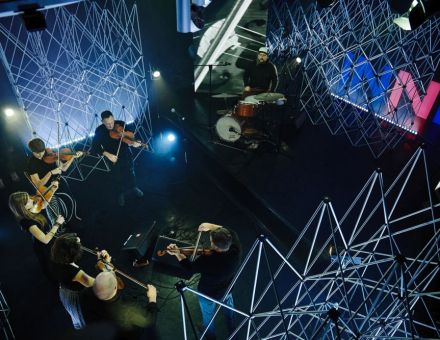 Rzut z góry.  W dole 7 instrumentalistów w studio Strefy Innych Brzmień. W  tle ledon z napisem INNE i aluminiowe kratownice.