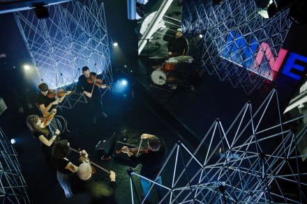 Rzut z góry.  W dole 7 instrumentalistów w studio Strefy Innych Brzmień. W  tle ledon z napisem INNE i aluminiowe kratownice.