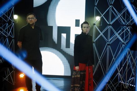 Zdjęcie zrobione zza aluminiowych kratownic. W kadrze kobieta i mężczyzna na tle świecącej ścianki led z logotypem festiwalu Inne Brzmienia.