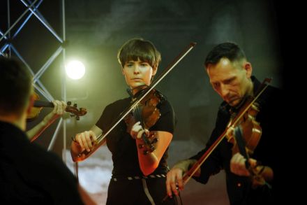 W centrum kadru młoda skrzypaczka w krótkich włosach, obok niej skrzypek .