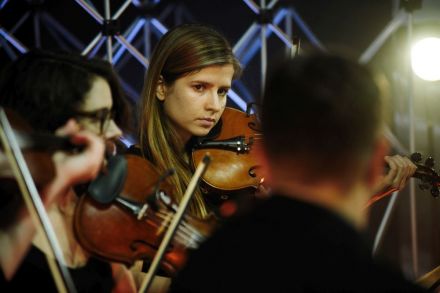 Grupa muzyków, w centrum kadru młoda skrzypaczka, blondynka. Wzrok skierowany w stronę aparatu.