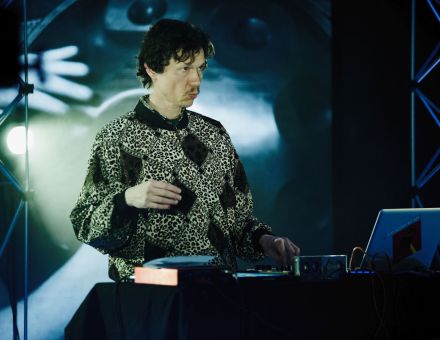 Mężczyzna za konsolą djską. W tle ekrany led z szarymi abstrakcyjnymi animacjami. Mężczyzna ma wąsik, jest ubrany w sweter w stylu lat 80-tych, rąby, panterka.