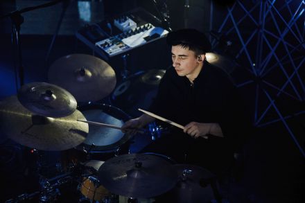 Zbliżenie na perkusistę. Ma zamknięte oczy, w rękach pałeczki, którymi gra na perkusji. W tle konsola. Mężczyzna jest ubrany na czarno, ma czarny beret.