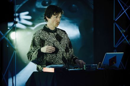 Mężczyzna za konsolą djską. W tle ekrany led z szarymi abstrakcyjnymi animacjami. Mężczyzna ma wąsik, jest ubrany w sweter w stylu lat 80-tych, rąby, panterka.