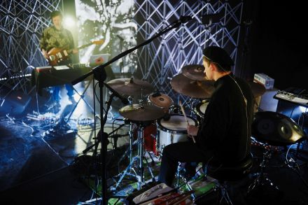 Szeroki plan, sala koncertowa, na pierwszym planie jest perkusista, siedzi  tyłem do obiektywu aparatu. Drugi plan to gitarzysta jest przodem do obiektywu. W tle ekrany ledz szarymi abstrakcyjnymi animacjami oraz aluminiowe kratownice. Dużo sprzętu elektronicznego: kable, mikrofony, konsole.