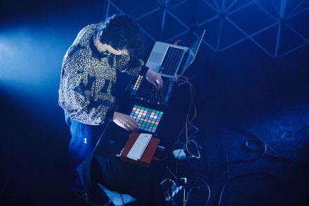 Zdjęcie z góry, dj przy konsoli. Mężczyzna jest ubrany w sweter w stylu lat 80-tych, rąby, panterka. Niebieskie światło.