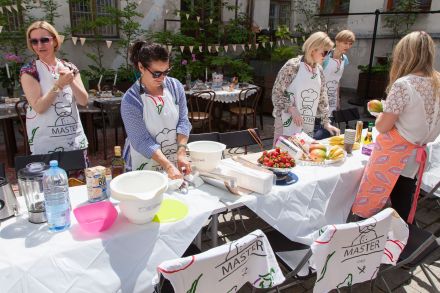 Na stole stoją naczynia, woda, owoce i blendery. Dookoła stołu stoją kobiety.