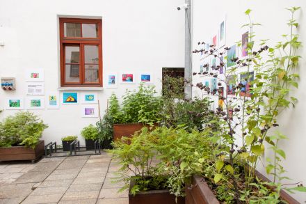 Zdjęcie z oddali całej wystawy. Kadr obejmuje dwie ściany patio, na których są zawieszone obrazy oraz stoją kwiaty.