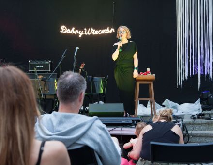 Zbliżenie na scenę, gdzie konferansjerka z zapowiada koncert.