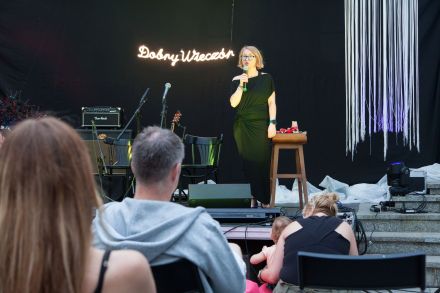 Zbliżenie na scenę, gdzie konferansjerka z zapowiada koncert.