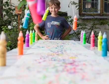 Zbliżenie na fragment stołu i kartki, na której powstały kolorowe mazaje. Dookoła kartki na stole stoją farby, a na końcu stołu stoi chłopiec i się uśmiecha.